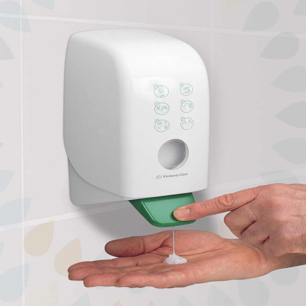 Eine Person spendet Kleenex® feuchtigkeitsspendende Hand- und Körperlotion (Kimberly-Clark GmbH) aus einer weißen 1-Liter-Wandkassette in einem gefliesten Badezimmer mit einem grünen Hebel für Hand- und Körperpflege. Packung: 6 Kassetten pro Karton.