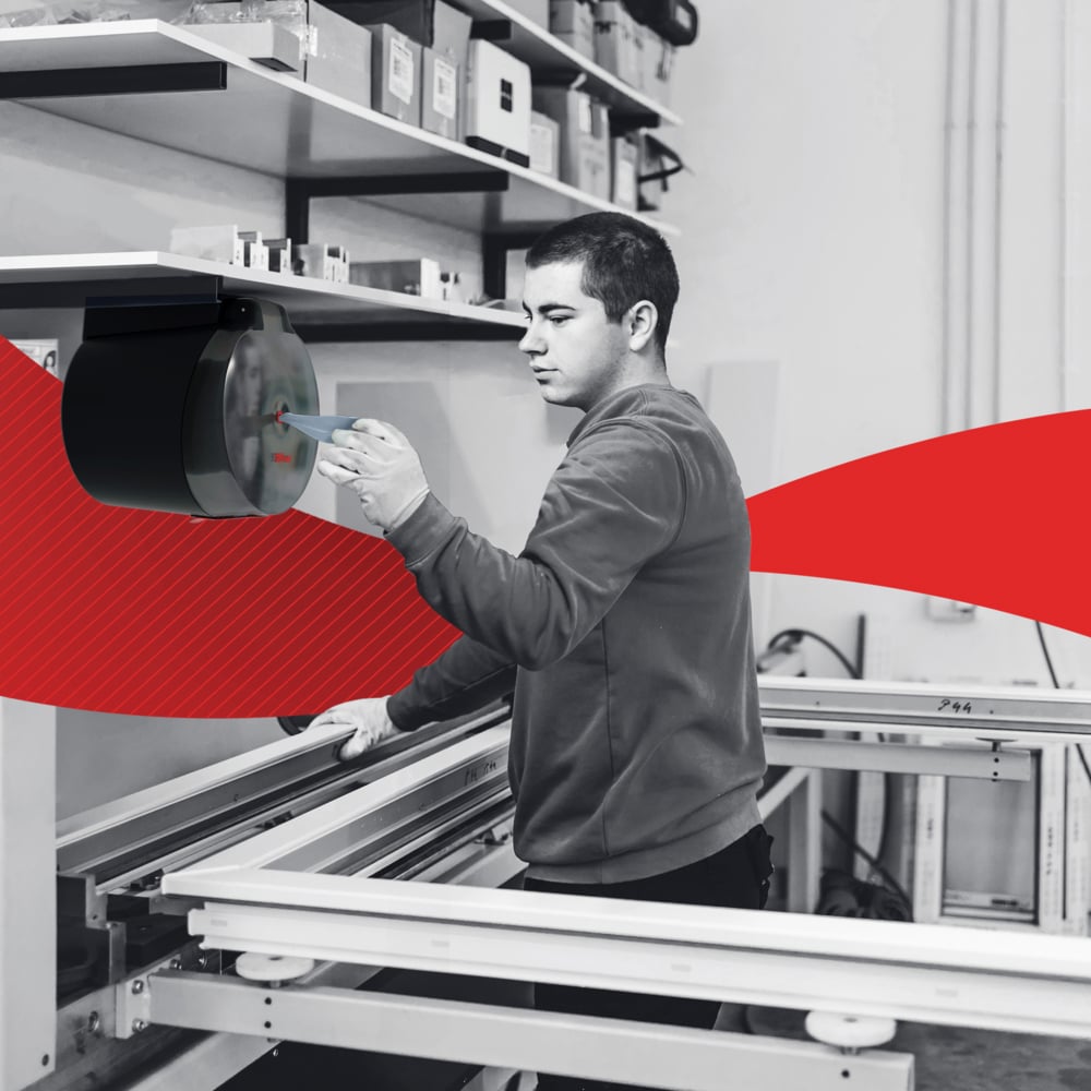 A man operates machines in a workshop. In the background are shelves with tools and the WypAll® L20 General Clean Centrefeed for Reach+™ (blue, 2-ply, 6x550 sheets) from Kimberly-Clark GmbH amidst red graphic elements.
