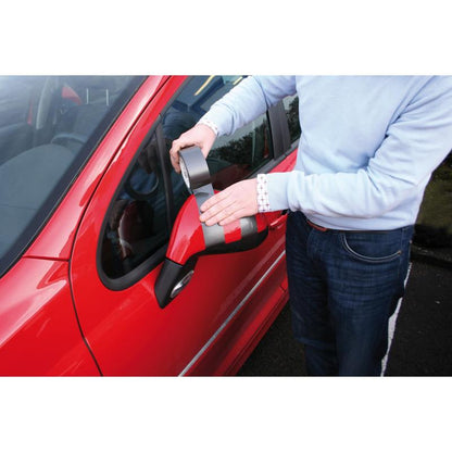 A person in a light blue sweater uses the Scotch® Extremium™ No Residue High Performance Duct Tape (18.2 m x 48 mm) from 3M to fix the damaged side mirror of a red car while standing at the driver's door. The gray tape is effective and leaves no residue.