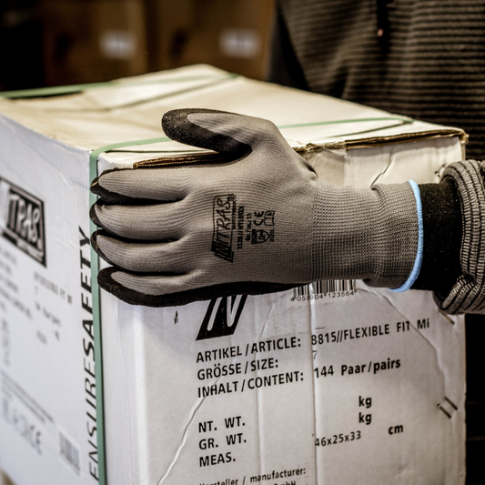 Eine Person, die Nitras Nylotex Nylonhandschuhe mit Latex-Beschichtung der AS Arbeitsschutz GmbH trägt, hält eine große weiße Schachtel mit deutscher und englischer Beschriftung in der Hand; nur ihr Oberkörper und ihr Arm sind teilweise sichtbar.