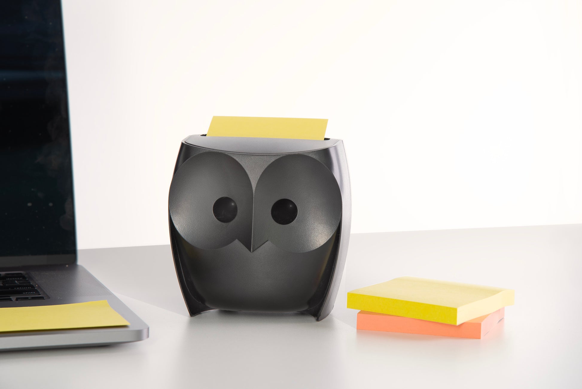 Ein schwarzer, eulenförmiger Post-it® Z-Notizenspender der 3M Deutschland GmbH hält eine gelbe Super Sticky Z-Note auf einem weißen Schreibtisch, neben einem Laptop und Stapeln von Post-it® Super Sticky Z-Notes in Gelb und Orange. Der Hintergrund ist hell und einfarbig.