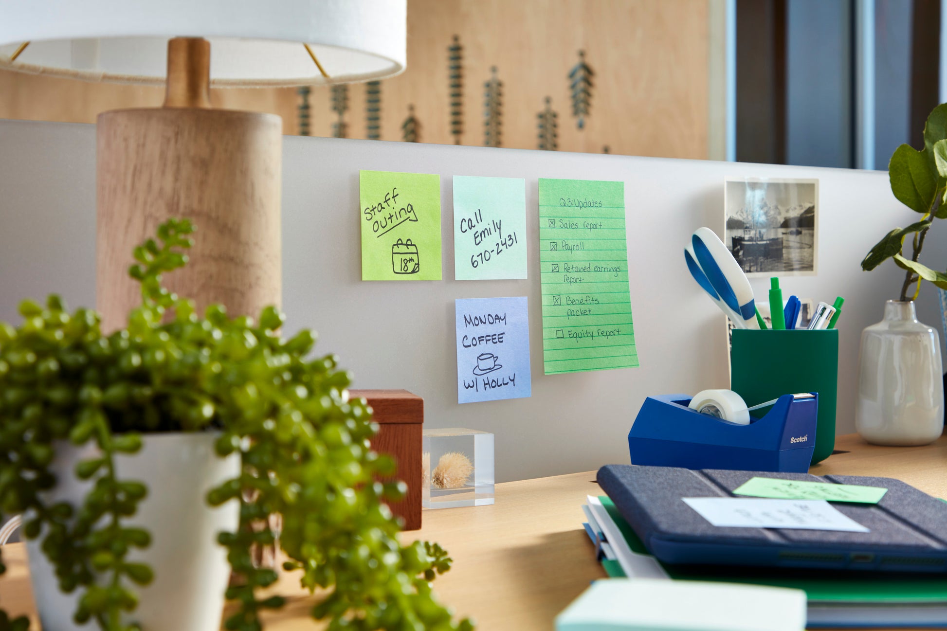 Auf einem Schreibtisch hängt ein 3M Post-it® Super Sticky Notes Block (101x101 mm, 70 Blatt, PEFC) der 3M Deutschland GmbH als Erinnerungshilfe an der Wand. Eine Lampe, eine Grünpflanze, eine Tasse mit Stiften, Büromaterial und ein Foto sind ebenfalls zu sehen.