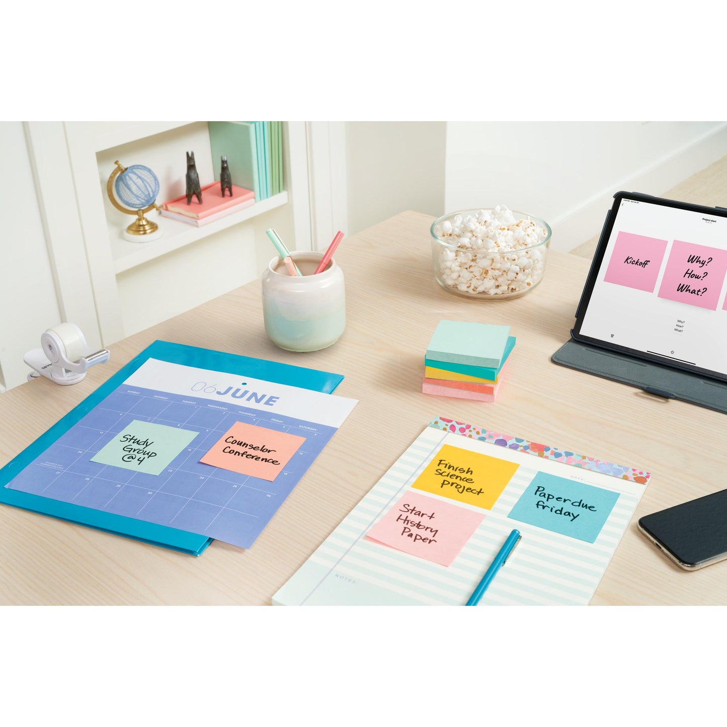 An organized desk for studying or working with Post-it® Notes (76 mm x 127 mm, 100 sheets/block, 6 blocks/pack, 100% PEFC) from 3M Deutschland GmbH, notebooks, pens in a cup, popcorn, tape, a tablet with notes; a bookshelf with books adorns the background.