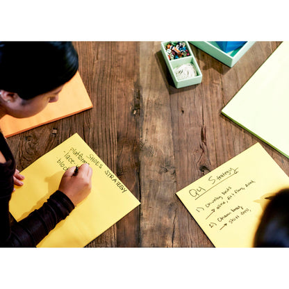 Two people sit at a wooden table writing on 3M Post-it® Super Sticky Big Notes BN11-EU, Yellow (279 x 279 mm, 30 sheets/block, 100% PEFC) and brainstorming ideas like "PHOTOSHOOT SHOES STRATEGY" and "Q4 Strategy". Yellow sticky notes and markers are ready.