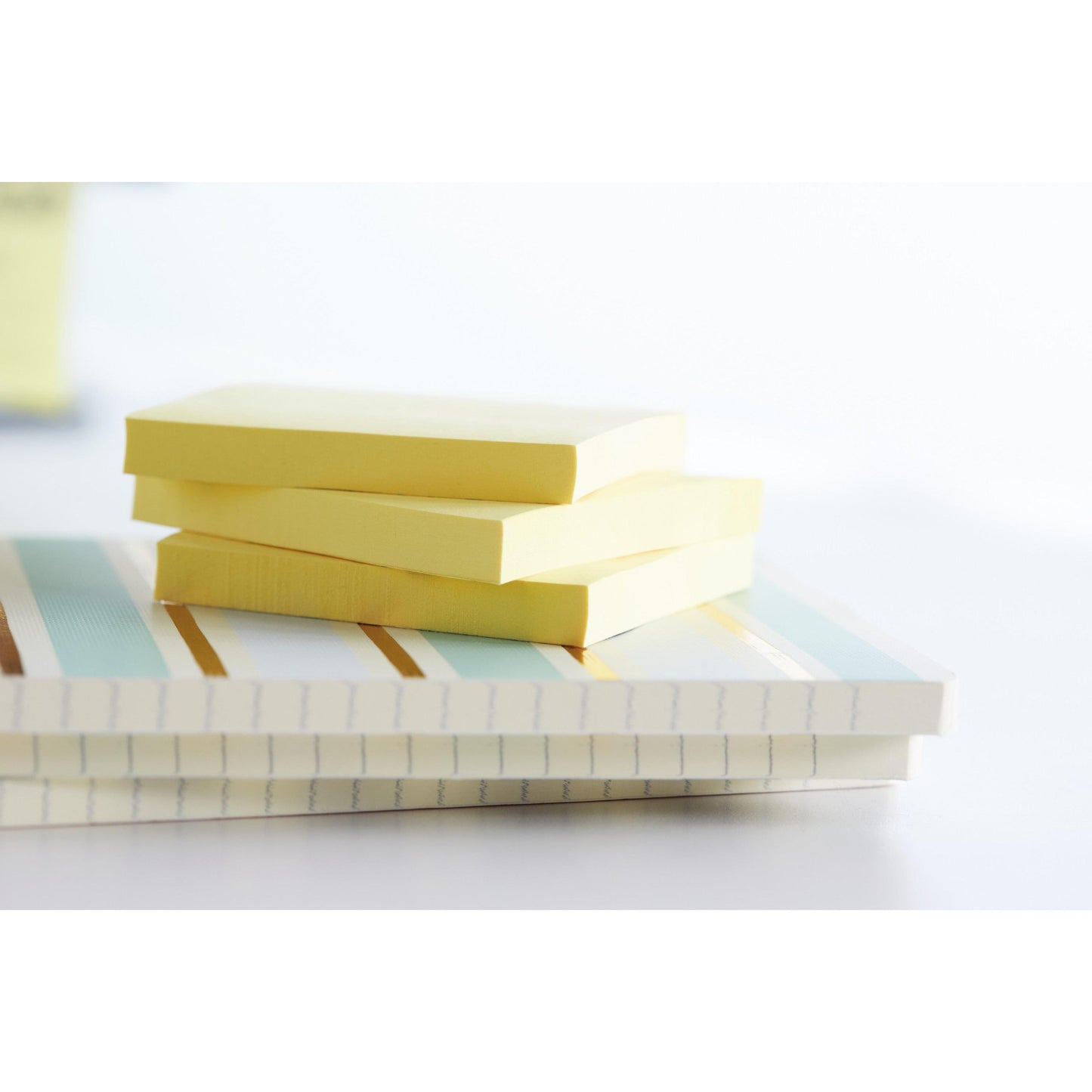 Three Post-it® Recycling Notes (76 mm x 76 mm, 100 sheets/block, 100% PEFC) from 3M Deutschland GmbH are stacked on two striped notebooks with white, blue, and gold lines, lying on a light surface with a blurred background.