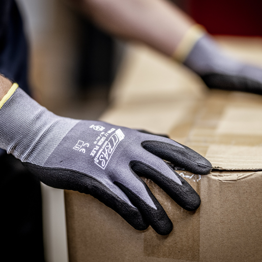 Eine Person trägt NITRAS SKIN FLEX Strickhandschuhe von AS Arbeitsschutz GmbH aus Spezial-Stretch-Strick, grau, teilbeschichtet, hebt einen versiegelten Karton. Sichtbar sind nur die behandschuhten Hände und ein Teil des Kartons.