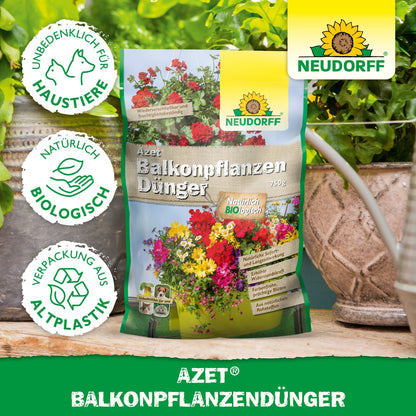 A bag of Azet Balcony Plant Fertilizer from Neudorff Shop stands on a table between potted plants and balcony boxes. The packaging shows flowers, text, and symbols for animal protection, organic ingredients, and packaging made from bioplastic.