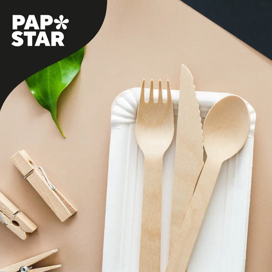 Ein Einwegteller aus Papier mit einer Holzgabel, einem Messer und einem Löffel steht auf einer beigen Fläche. Ein grünes Blatt, zwei hölzerne Wäscheklammern und das Papstar-Logo sind ebenfalls auf dem Bild zu sehen.