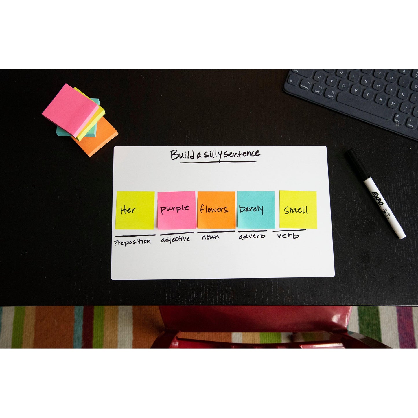 On a whiteboard are five colorful Post-it® Notes (76 mm x 127 mm, 100 sheets/block, 6 blocks/pack, 3M Deutschland GmbH) labeled with words and parts of speech; nearby are sticky notes, a marker, a keyboard, and a striped carpet.