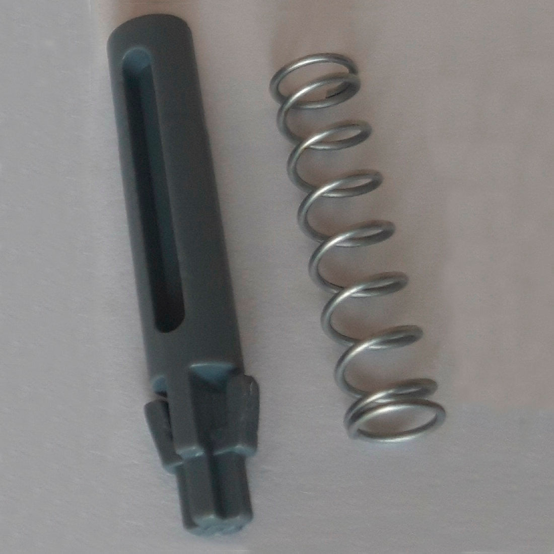 A metal spring and a dark grey cylindrical replacement door pin with grooves and notches, part of the DI Protect door pin & spring (1 piece) from Diversey Deutschland GmbH & Co. OHG, are depicted on a light surface and showcase the robust materials for the DI Protect trolley.