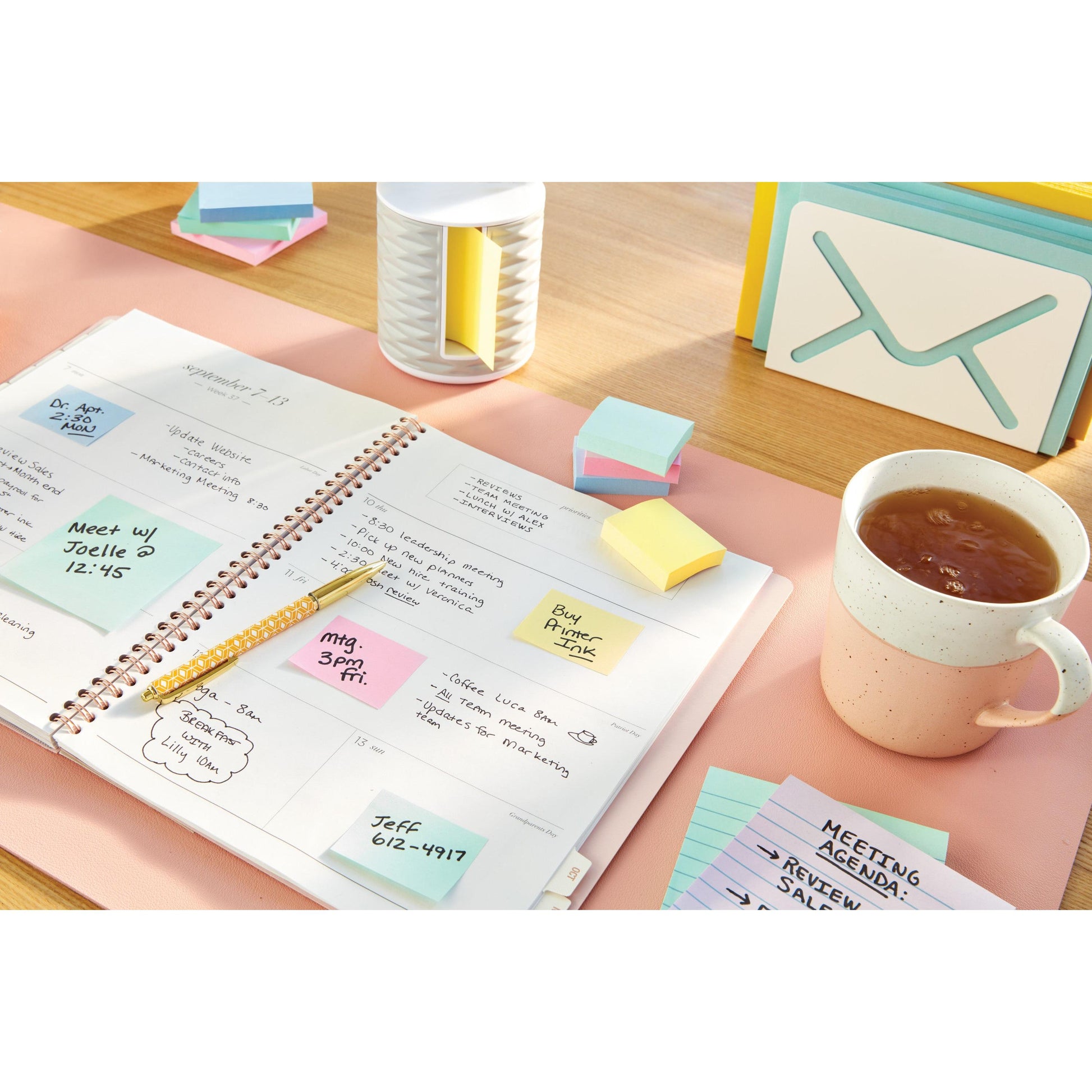 Ein aufgeschlagener Spiralplaner liegt auf einem Schreibtisch, umgeben von 3M Deutschland GmbH Post-it® Notes (38 mm x 51 mm, Gelb), einem goldenen Stift, einer Tasse Tee, einem Kartenhalter in Form eines Umschlags und "MEETING AGENDA"-Karten.