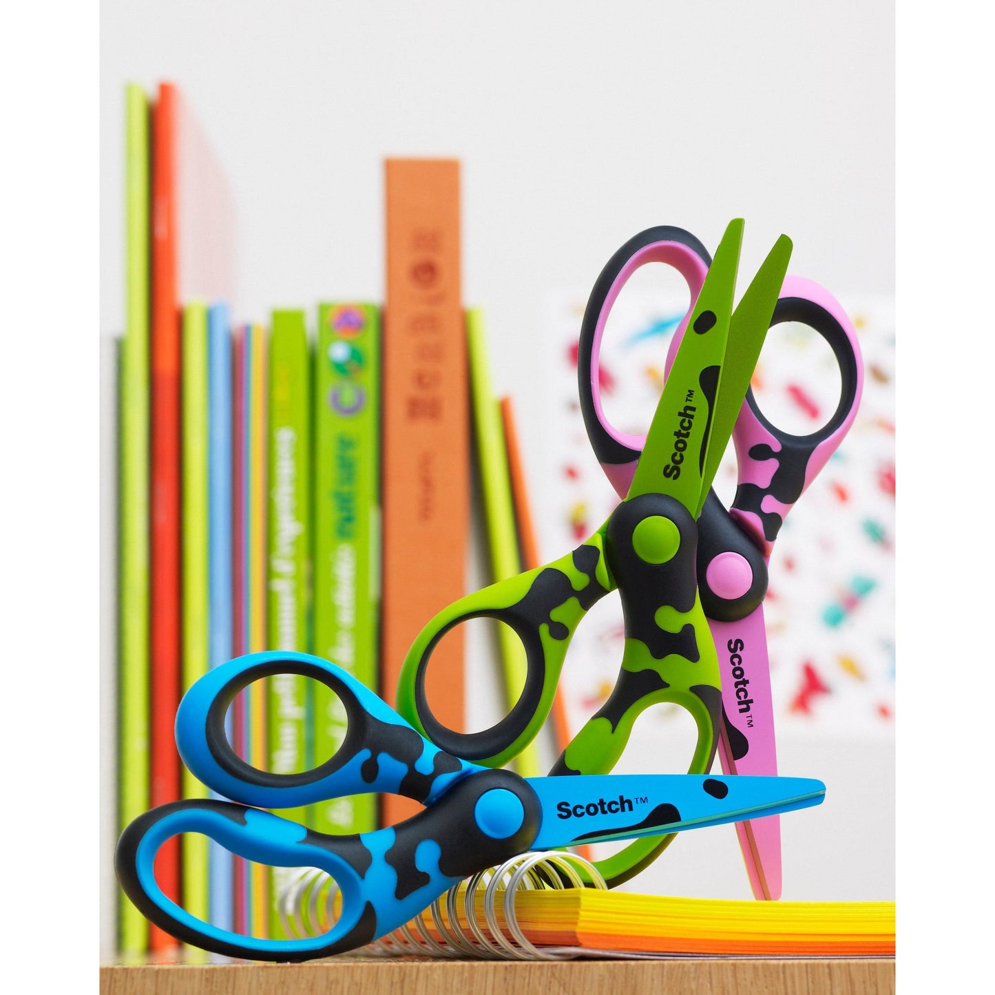 Two Scotch™ DECO children's scissors (green, blue, or pink, 13 cm) from 3M Deutschland GmbH with colorful patterns and rounded tips lie on a stack of paper. In the background, there are colorful books.