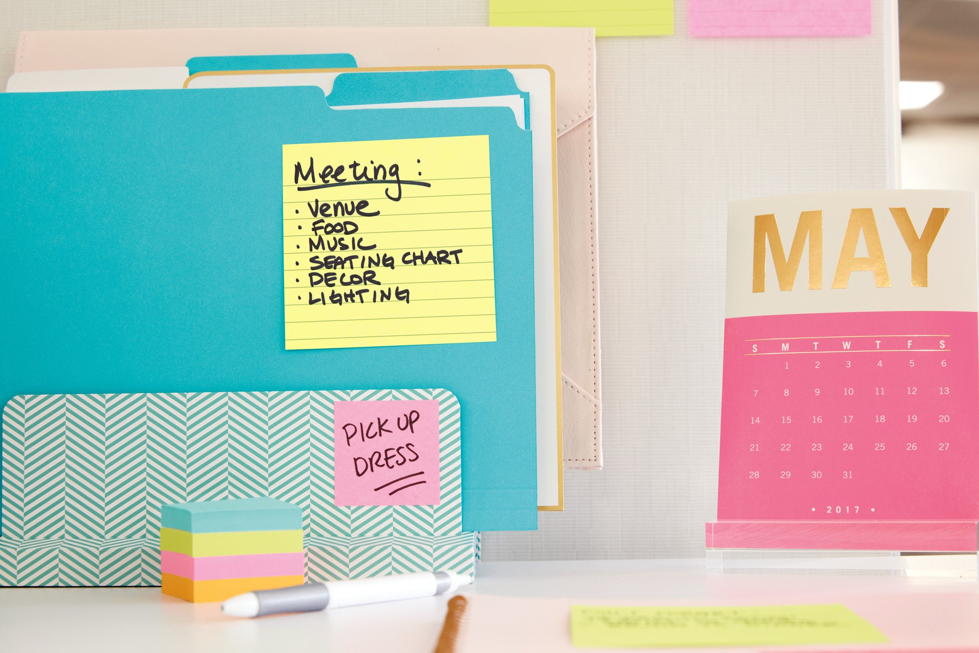 Ein Schreibtisch mit bunten Ordnern, einem Stapel Haftnotizen und einem rosa-goldenen Kalender für Mai 2017. Auf den Post-it® Super Sticky Notes (101 mm x 101 mm, 90 Blatt, PEFC) der 3M Deutschland GmbH sind Besprechungsthemen und "PICK UP DRESS.