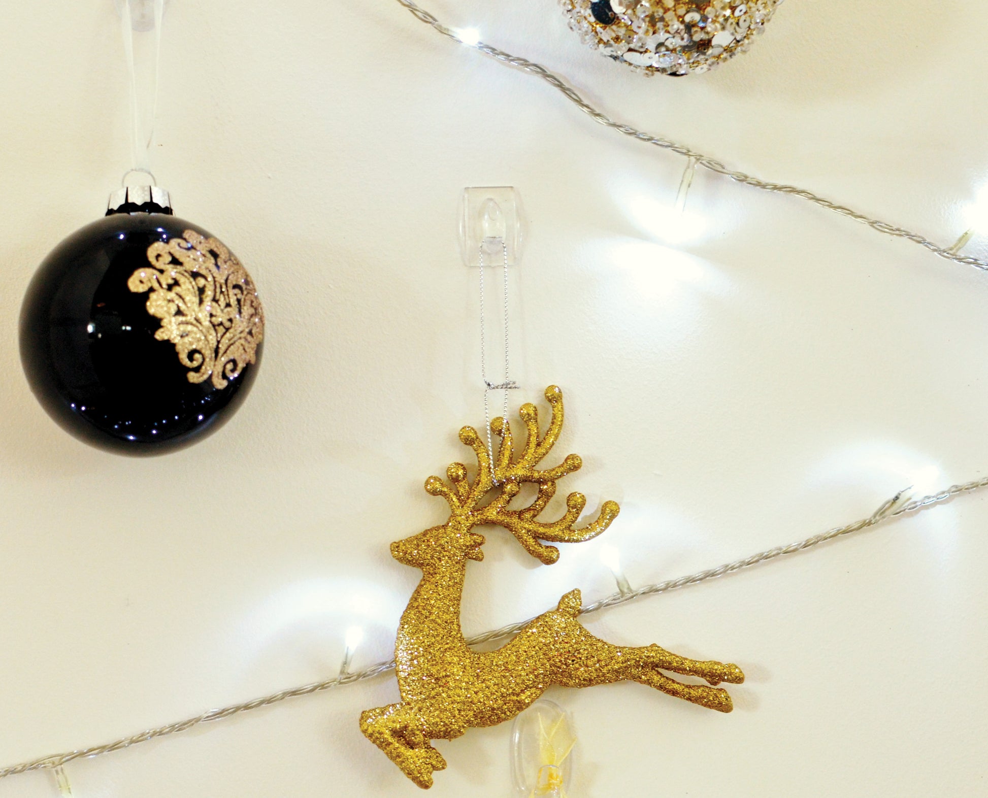 Ein goldglitzerndes Rentierornament wird mit einem transparenten Command™ Mini-Haken der 3M Deutschland GmbH an die Wand gehängt und neben schwarz-goldenen Weihnachtskugeln und weißen Lichterketten präsentiert.