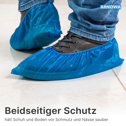 Nahaufnahme einer Person mit ARNOMED Xtra Strong CPE Schuhüberziehern von ARNOWA GmbH über dunklen Schuhen und Jeans. Die wasserdichten Einweg-Überzieher schützen Schuhe und Böden zuverlässig vor Schmutz und Feuchtigkeit. Beutel à 100 Stück.