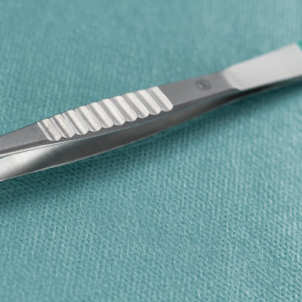 A close-up of a Paul Hartmann AG Hartmann splinter forceps straight 9cm, sterile (pack of 40 pieces), lying next to a metal surgical scalpel on a teal-colored, textured medical cloth.