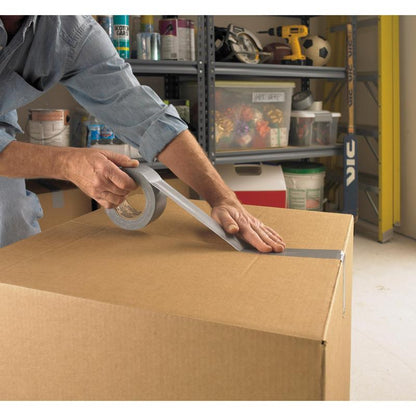 A person seals a large box with Scotch® Extremium™ Universal Adhesive Tape (3 m x 19 mm) from 3M Deutschland GmbH in a garage. In the background, shelves with tools, paint cans, and other items are visible.