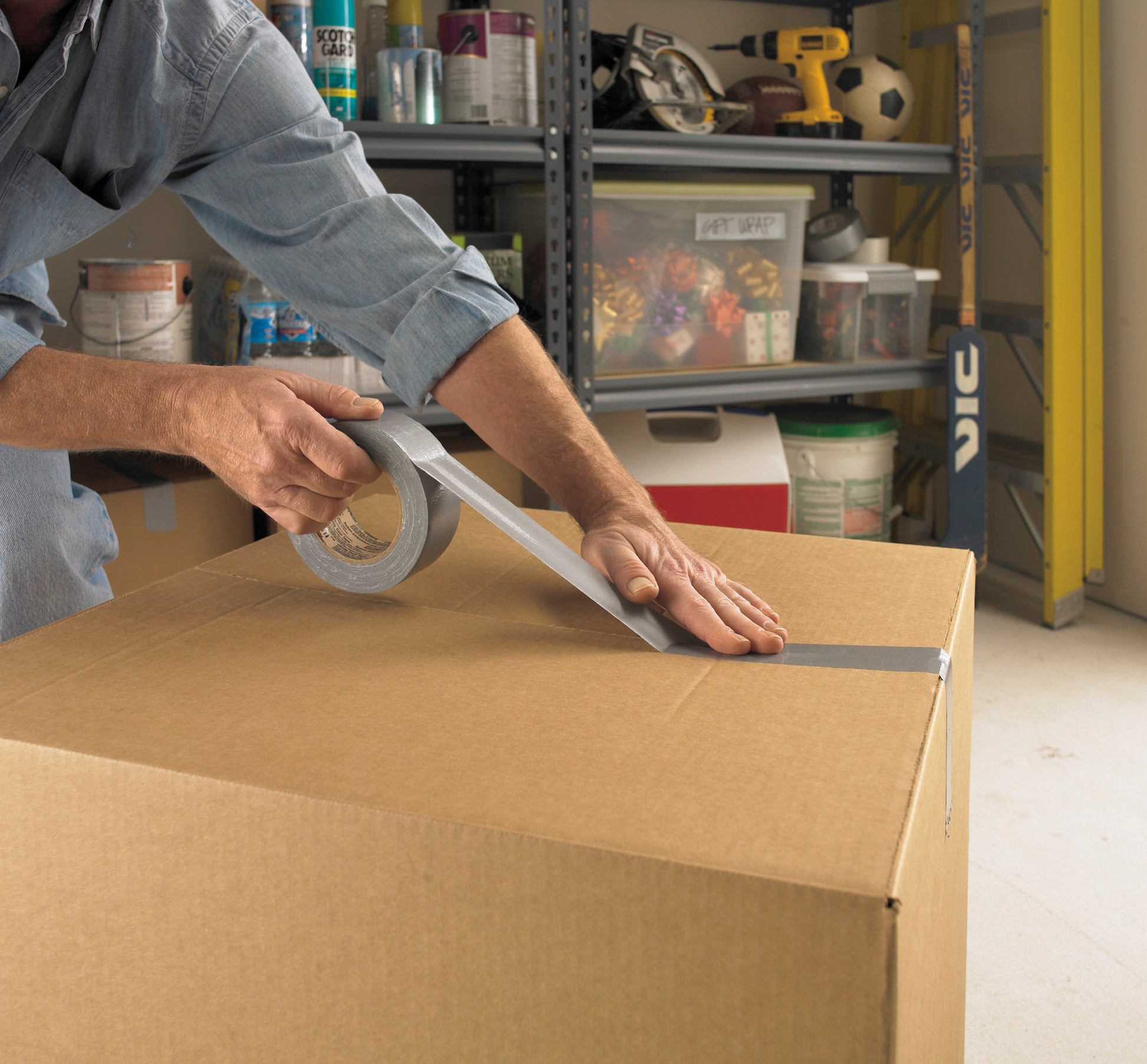 Eine Person verschließt einen großen Karton in einer Garage mit Scotch® Universal Klebeband 2904 (50 m x 48 mm) von 3M Deutschland GmbH, im Hintergrund sind Regale mit Werkzeug und Zubehör zu sehen.