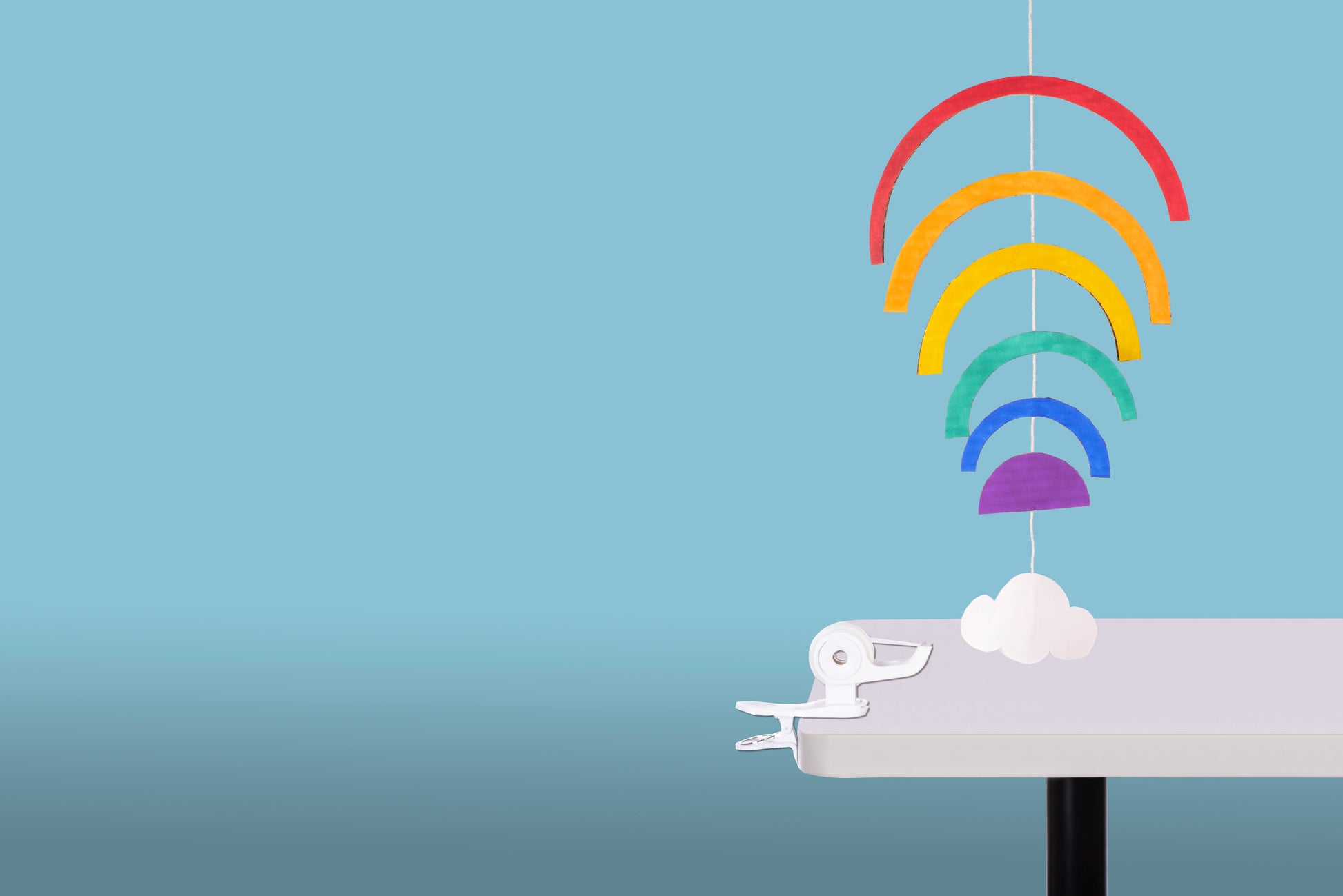 Ein farbenfrohes Papiermobile mit fünf Regenbogenbögen und einer weißen Wolke hängt an der Kante eines weißen Tisches, gesichert durch den 3M Deutschland GmbH Scotch™ Clip & Twist Klebeband-Abroller C19 mit Magic™ Tape, vor einem hellblauen Hintergrund.