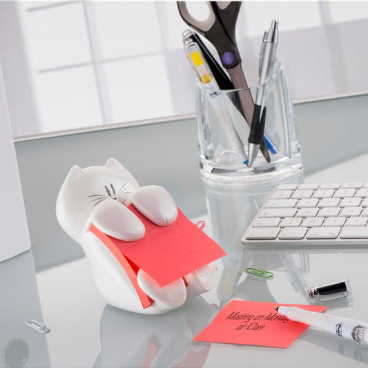 Ein weißer, katzenförmiger Post-it® Z-Notes Dispenser von 3M hält rosa Super Sticky Z-Notes (76x76mm) auf einem Glastisch mit einer roten Notiz in der Nähe, einer Tastatur, losen Büroklammern und einem durchsichtigen Becher mit Scheren und Stiften.
