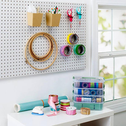 Ein Bastelarbeitsplatz mit einer Stecktafel, an der die Scotch™ Präzisionsschere rot von 3M Deutschland GmbH, Stickrahmen, Klebebandrollen und Stifte hängen. Unten sind Bastelmaterial und Bänder auf einem weißen Tisch in der Nähe eines Fensters angeordnet.