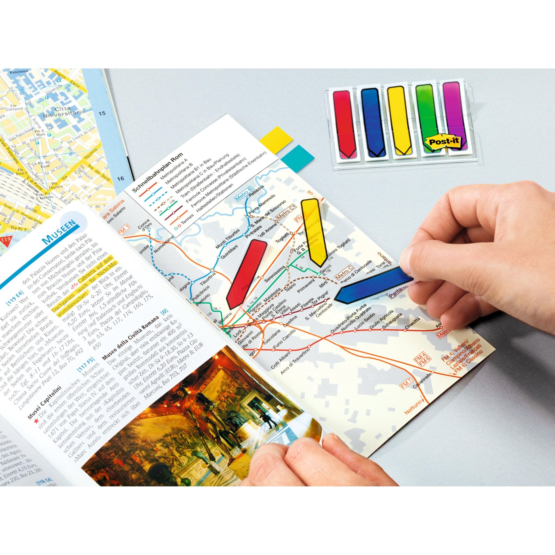 Eine Person markiert einen Stadtplan in einem Reiseführer mit einem blauen 3M Post-it® Index Pfeil (11,9 mm x 43,2 mm) aus dem 100er-Spender; andere farbige Pfeiltabs aus der gleichen Packung sind ebenfalls auf dem Tisch zu sehen.