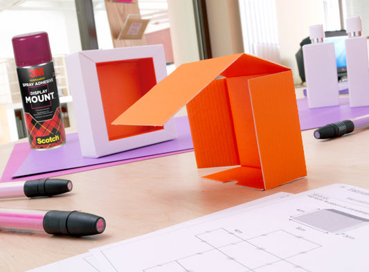 Ein orangefarbenes Architekturmodell aus Pappe steht auf einem Schreibtisch mit Zeichnungen, Markern, weißen Flaschen und einer Dose 3M™ Scotch® DisplayMount™ Klebespray (400 ml, 1 Dose/Packung) der 3M Deutschland GmbH im Hintergrund.
