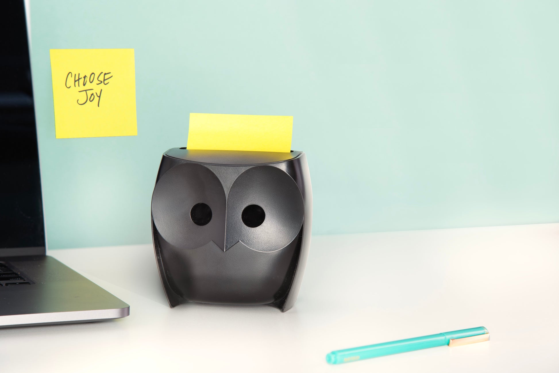 Ein 3M Post-it® Z-Zettelspender im schwarzen Eulendesign mit Super Sticky Z-Zetteln liegt auf einem weißen Schreibtisch neben einem türkisfarbenen Stift und einem Laptop, auf dem ein gelber Zettel mit der Aufschrift "CHOOSE JOY.