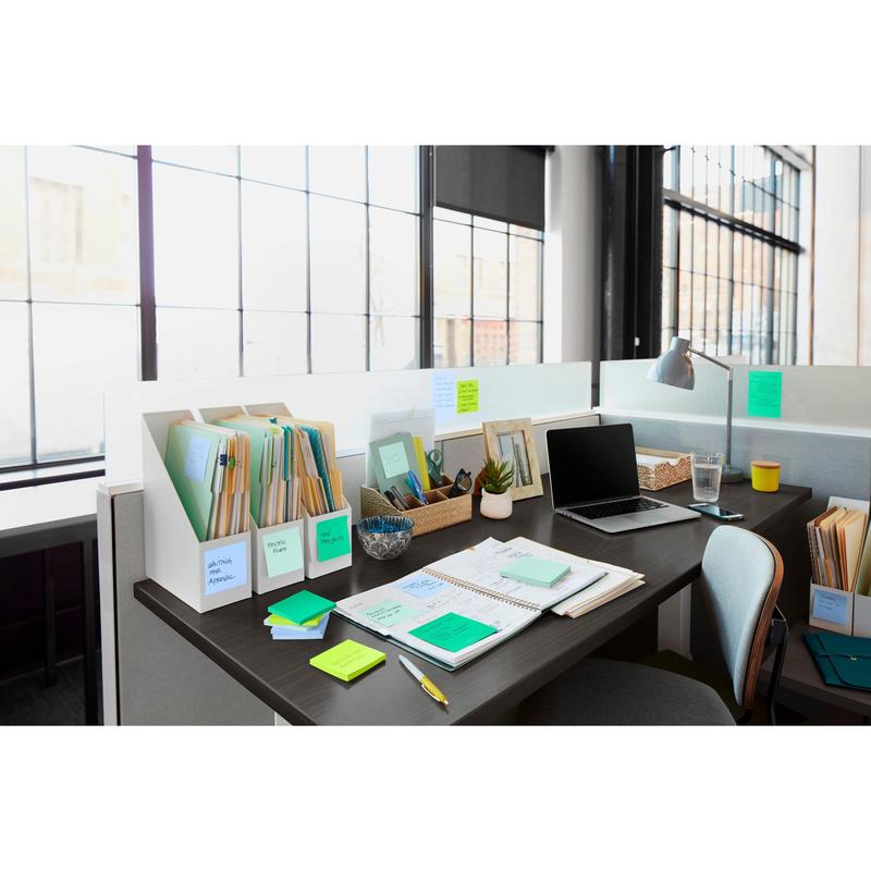 A modern office desk with Post-it® Super Sticky Notes (76 mm x 76 mm, 90 sheets/block, 100% PEFC) from 3M Deutschland GmbH, folders, notebooks, a laptop, a lamp, a pen holder, a small plant, and a glass of water in front of sunlit windows.