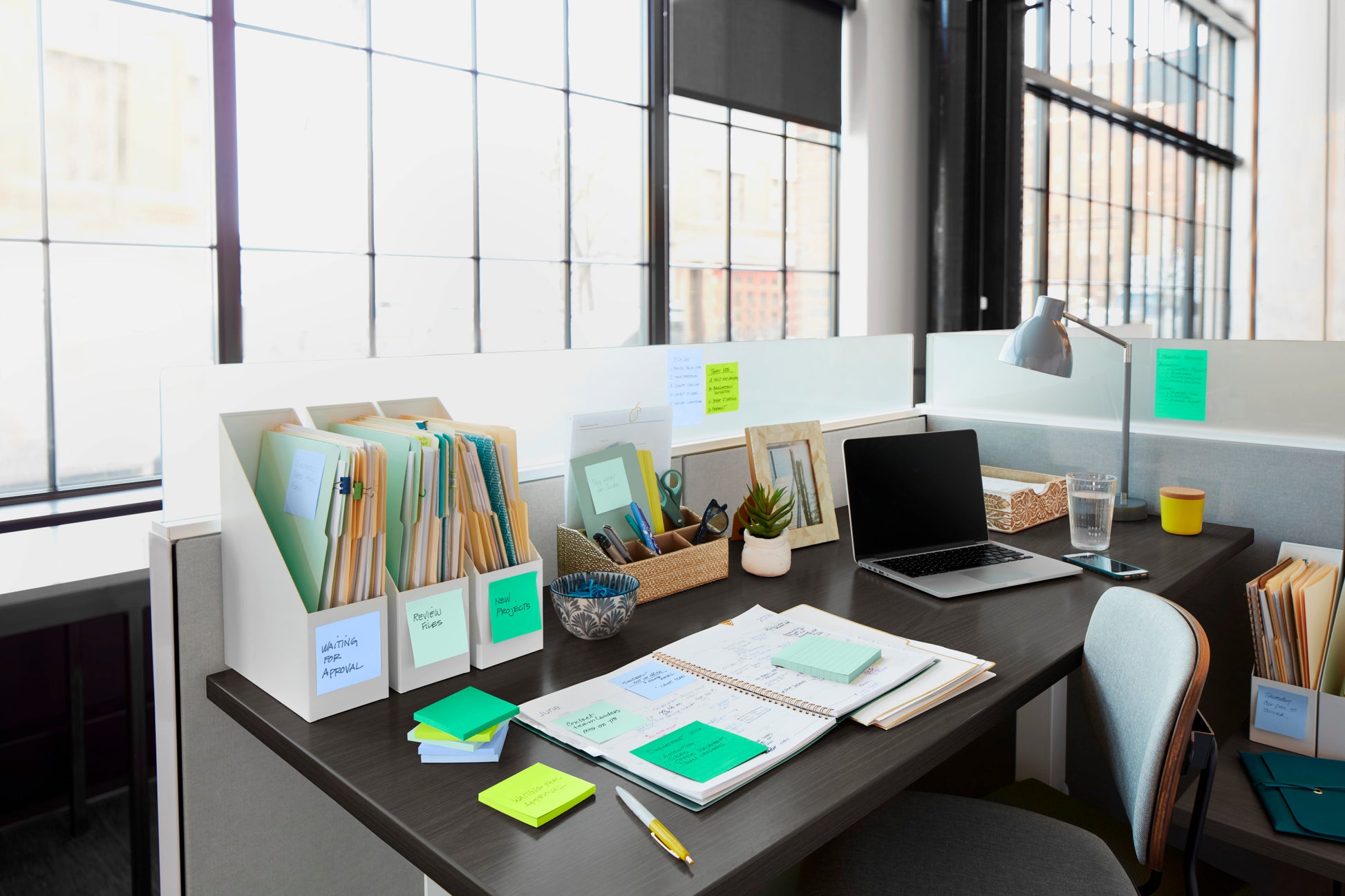Ein moderner Büroarbeitsplatz mit einem aufgeräumten Schreibtisch, Laptop, Notebooks, Ordnern, einem Wasserglas, einer Pflanze und einer Lampe. Auf dem Schreibtisch liegen 3M Deutschland GmbH Post-it® Super Sticky Notes im Großformat (101 mm x 101 mm). Große Fenster lassen natürliches Licht herein.