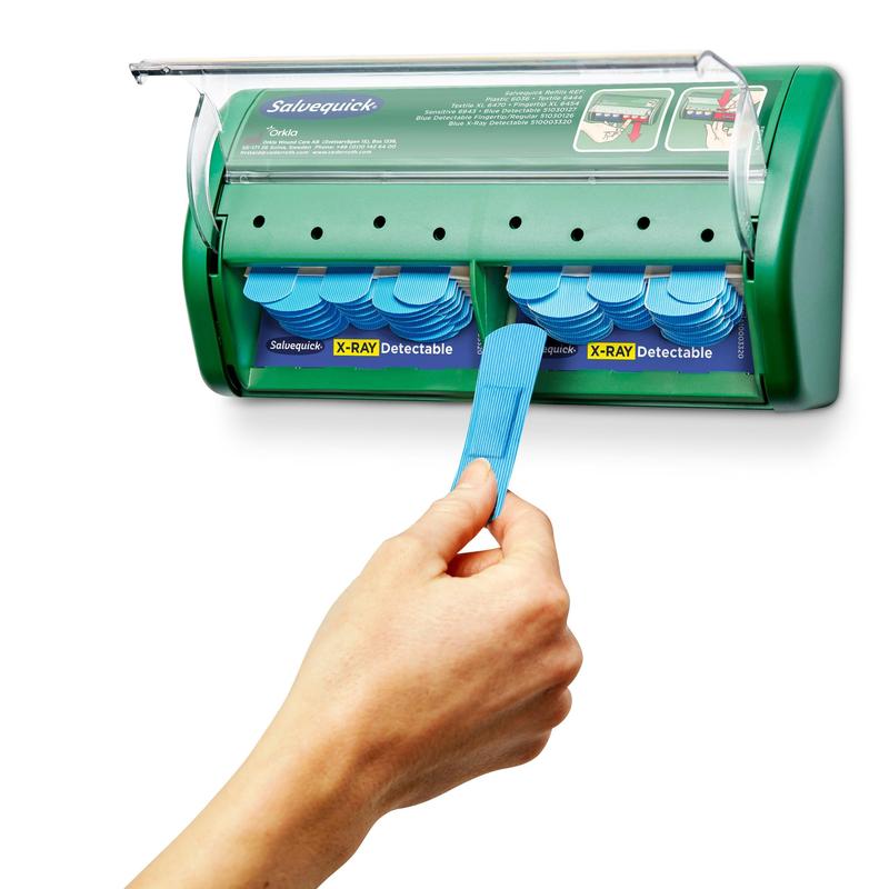 Eine Hand zieht einen Verband aus einem grünen Salvequick röntgendetektierbarer Pflasterspender (Orkla Wound Care AB), der 2x30 Gipsnachfüllungen enthält, wobei zusätzliche Verbände im Inneren des Spenders sichtbar sind.