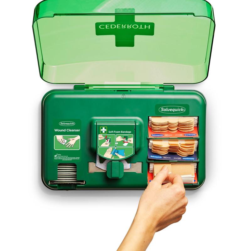 Eine Hand zieht einen Verband aus dem grünen Cederroth Wound Care Dispenser (20,3x30,6x15,5 cm) von Orkla Wound Care AB, der Wundreiniger und verschiedene Verbände in Fächern aufbewahrt.
