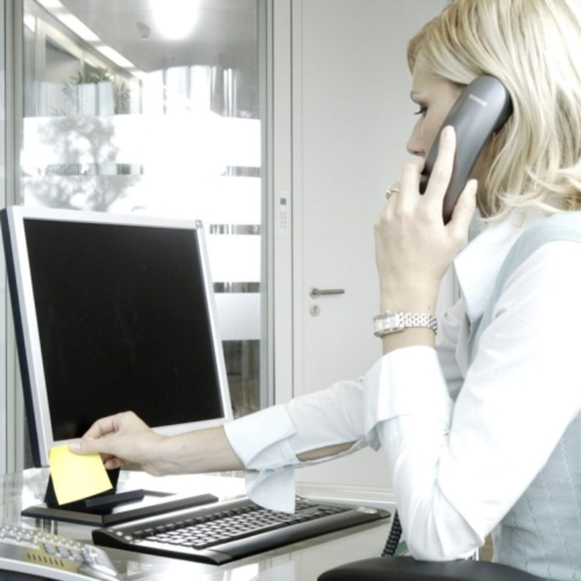 Eine blondhaarige Person an einem Schreibtisch legt einen 3M Post-it® Notes Würfel 636-B, Gelb (76x76 mm, 450 Blatt) auf ihren Monitor, während sie telefoniert; eine Tastatur und eine Maus befinden sich ebenfalls auf dem Schreibtisch.
