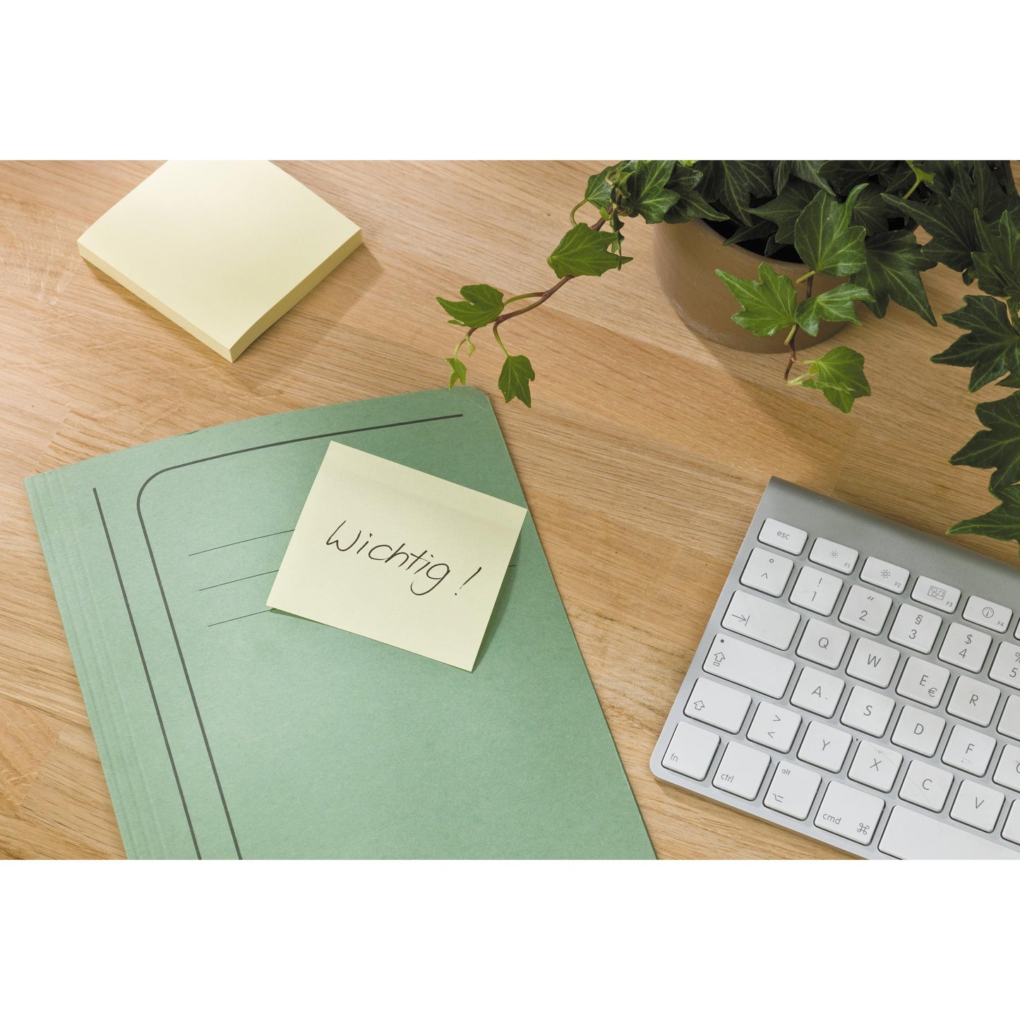 A green folder with a Post-it® Recycling Z-Note (76x76 mm, 100 sheets, 100% PEFC) by 3M Deutschland GmbH with the inscription "Important!" lies on a wooden table next to a keyboard, an ivy pot, and other sticky notes.
