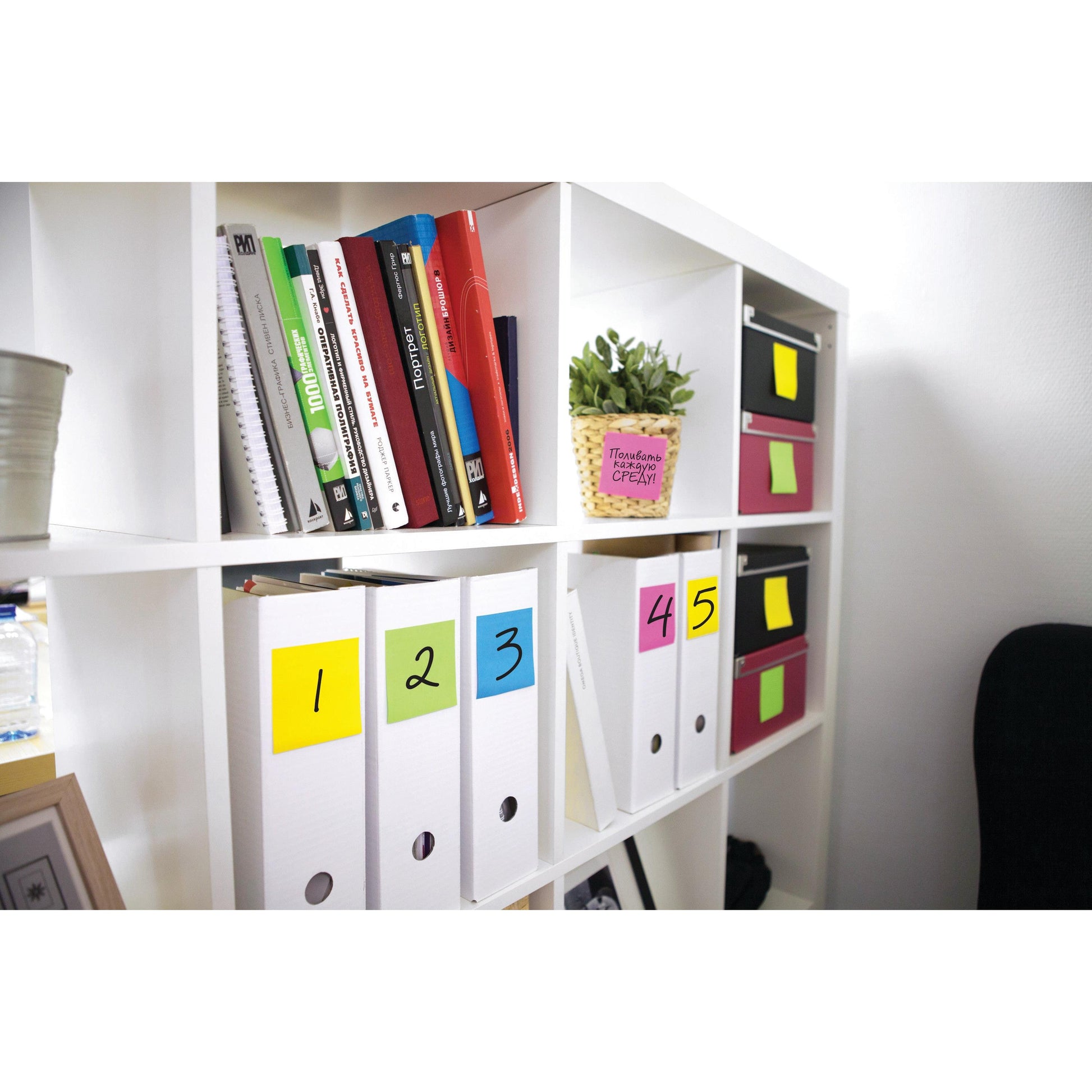 Ein weißes Regal enthält Bücher, Ordner 1-5, eine Topfpflanze in einem rosafarbenen Behälter und Aufbewahrungsboxen, die mit Post-it® Notes Würfel 636-B, Gelb von 3M Deutschland GmbH (76x76mm, 450 Blatt pro Packung) beschriftet sind, sowie andere Büroartikel.