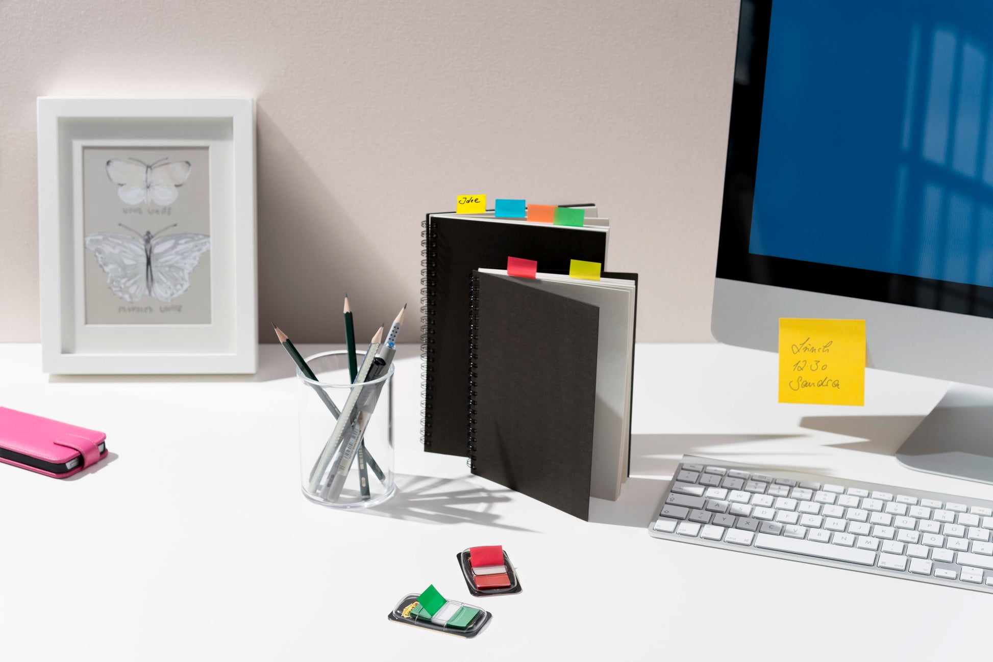 Ein aufgeräumter Schreibtisch zeigt Notizbücher mit Post-it® Index Haftstreifen der 3M Deutschland GmbH, Bleistifte in einem Glas, einen gerahmten Schmetterling, einen Computerbildschirm mit Haftnotiz, Tastatur, Radiergummis und ein rosa Mäppchen auf einer weißen Fläche.