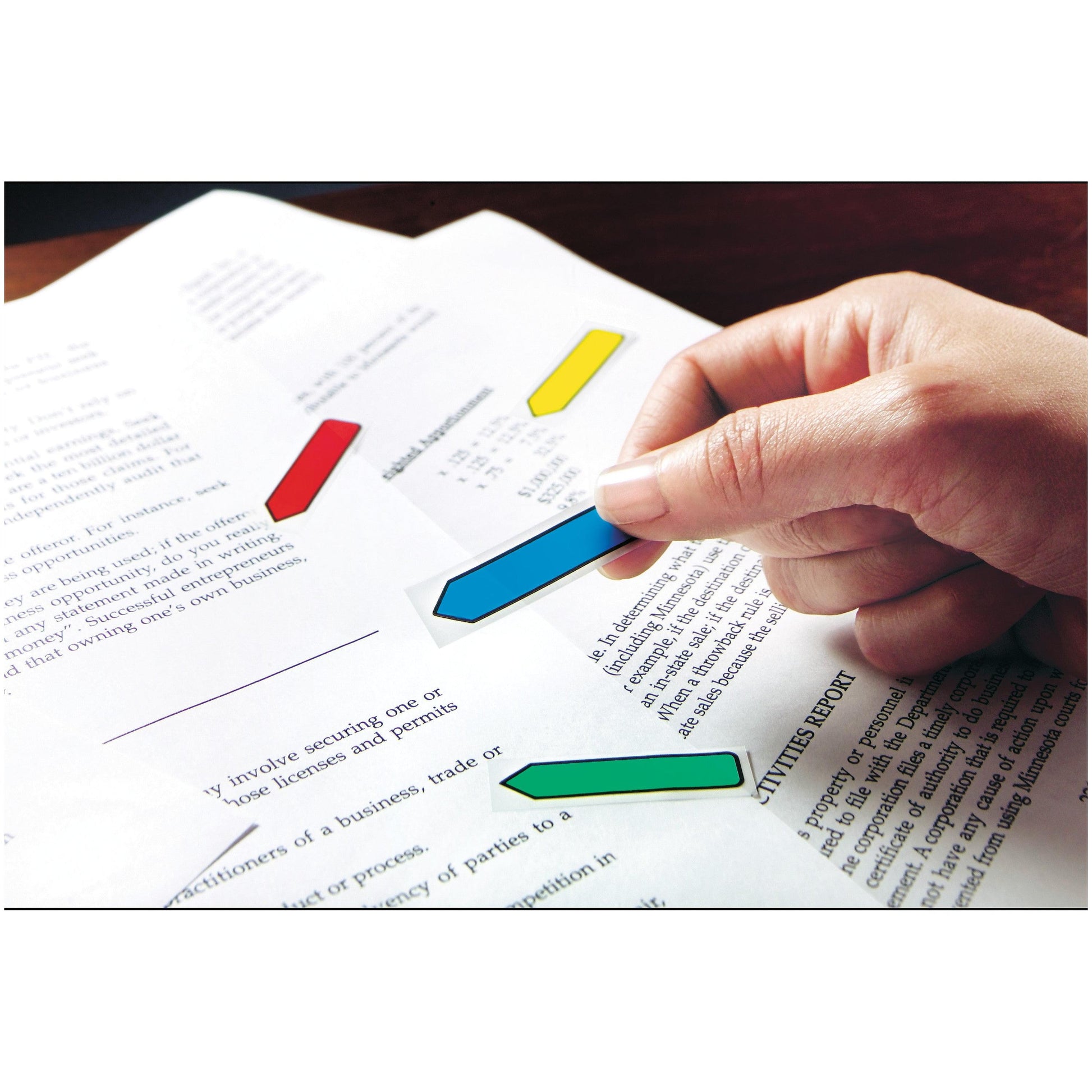 Eine Hand klebt einen grünen Post-it® Indexpfeil (11,9 mm x 43,2 mm) der 3M Deutschland GmbH auf ein Dokument, das bereits mit blauen, roten und gelben Tabs markiert ist; der Text des Dokuments ist teilweise sichtbar.
