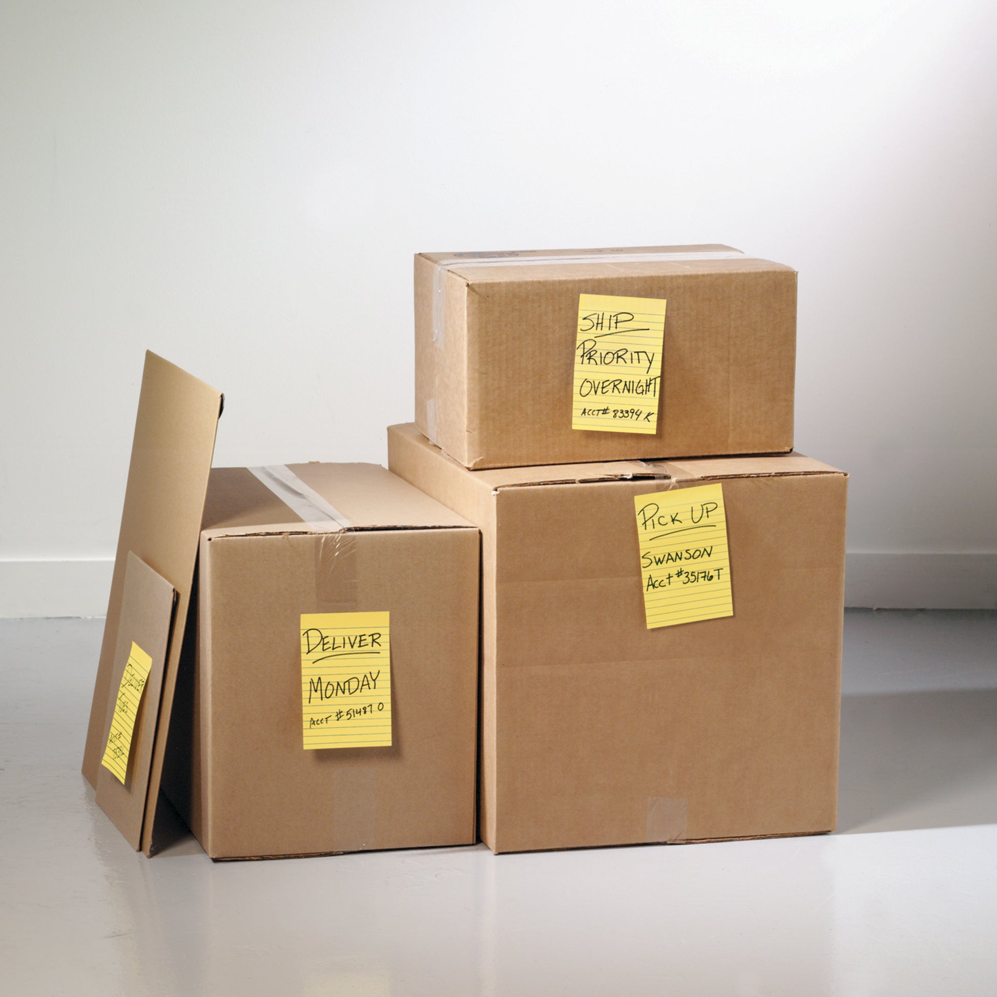 Vier in Innenräumen gestapelte Kartons mit je einer 3M Post-it® Super Sticky Note in Ultra Gelb (101×152 mm) von 3M Deutschland GmbH mit der Aufschrift "Deliver Monday". Einfarbiger Wandhintergrund; die Zettel sind 100% PEFC-zertifiziert, SGSCH-PEFC-COC-110078.