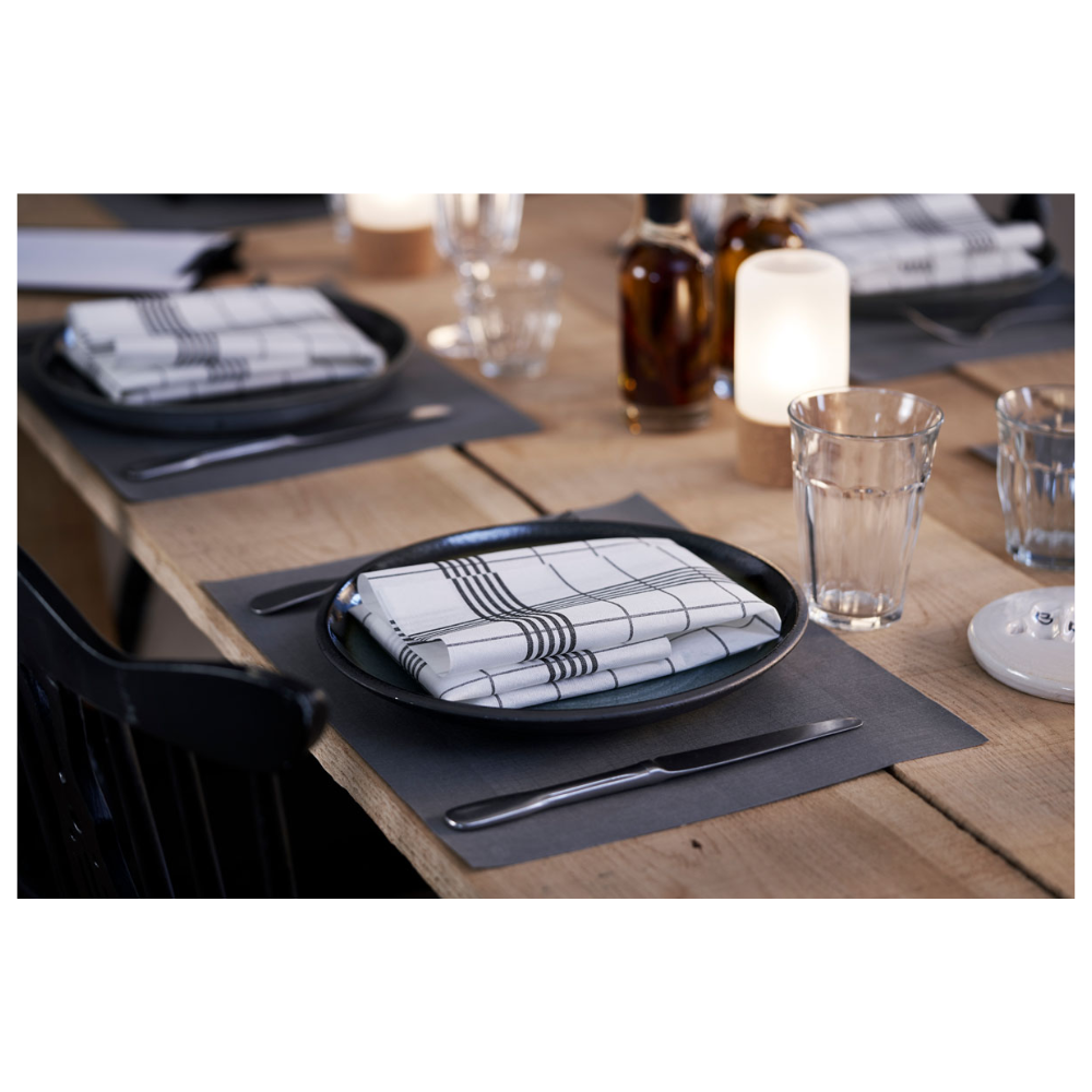 A dining table set consists of plates in Linnea Black by Duni GmbH and checkered napkins. In the background, three glass bottles stand next to burning candles. Water glasses and cutlery are elegantly presented on a Duni Dunicel® placemat 30 x 40 cm, with the light wooden table emphasizing the simple elegance.