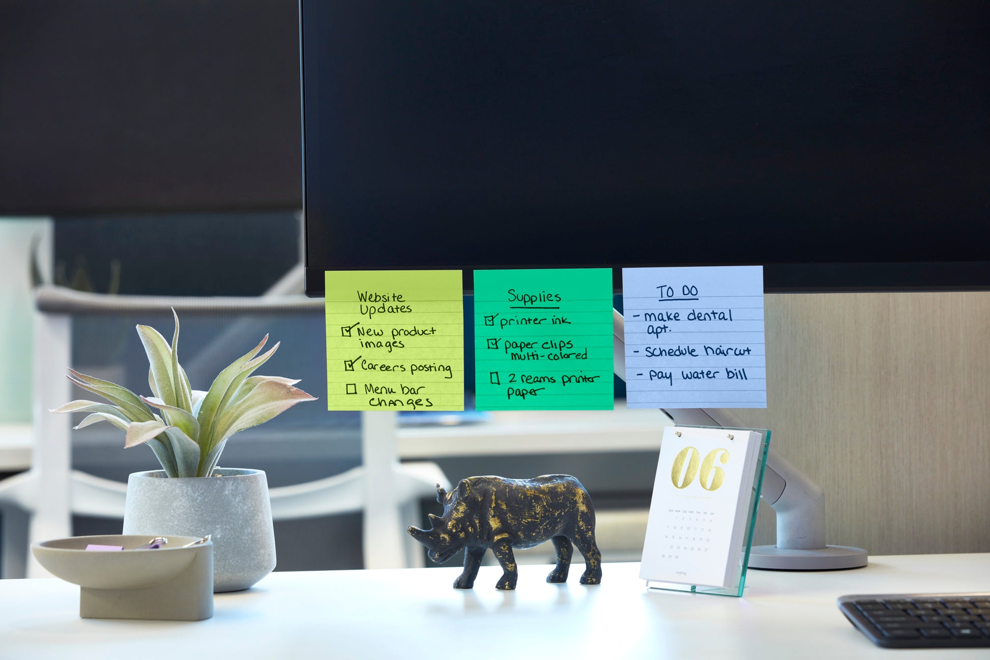 Ein Computermonitor mit Post-it® Super Sticky Notes (101 mm x 101 mm, 90 Blatt, 100% PEFC) der 3M Deutschland GmbH, auf denen Aktualisierungen der Website und zu erledigende Aufgaben aufgelistet sind. In der Nähe: eine Topfpflanze, eine Spitzmaulnashorn-Figur, ein Tischkalender mit dem Datum "06" und eine Schale mit Büroklammern.