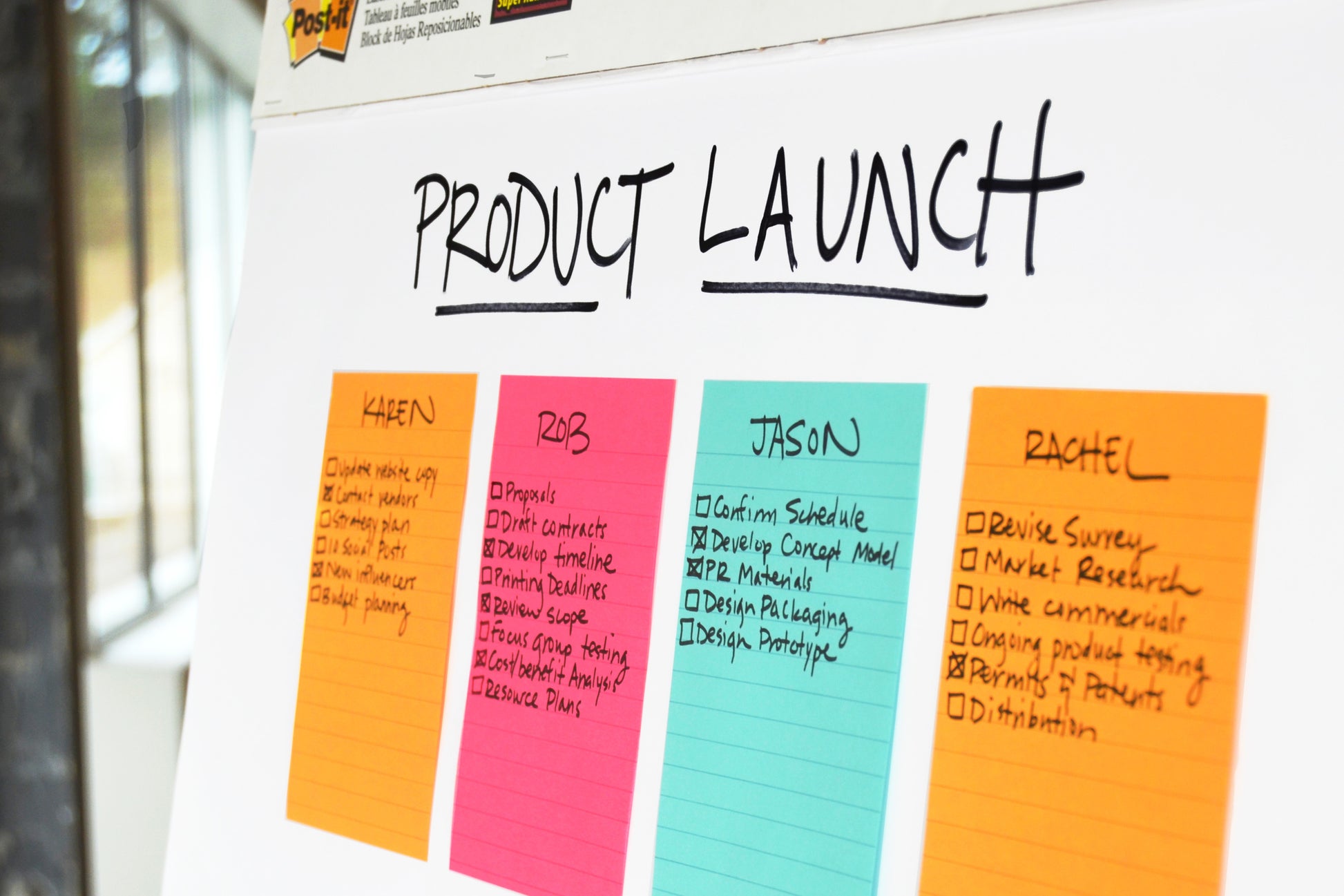 Auf einem Whiteboard mit der Aufschrift "PRODUCT LAUNCH" stehen 3M Deutschland GmbH Post-it® Super Sticky Notes im Großformat (101 mm x 101 mm, 70 Blatt/Block, 100% PEFC), jeweils beschriftet mit Karen, Rob, Jason und Rachel mit Aufgaben-Kästchen in einem hellen Arbeitsbereich.