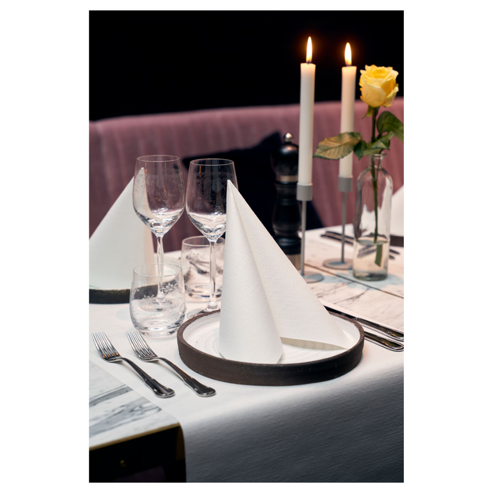 A dining table is elegantly set with two Duni Dunilin® napkins 48 x 48 cm White, folded into triangles on plates. Two large burning candles and a yellow rose in a glass vase adorn the background. Wine glasses, a water glass, and cutlery complete this sophisticated ambiance with fabric-like charm.
