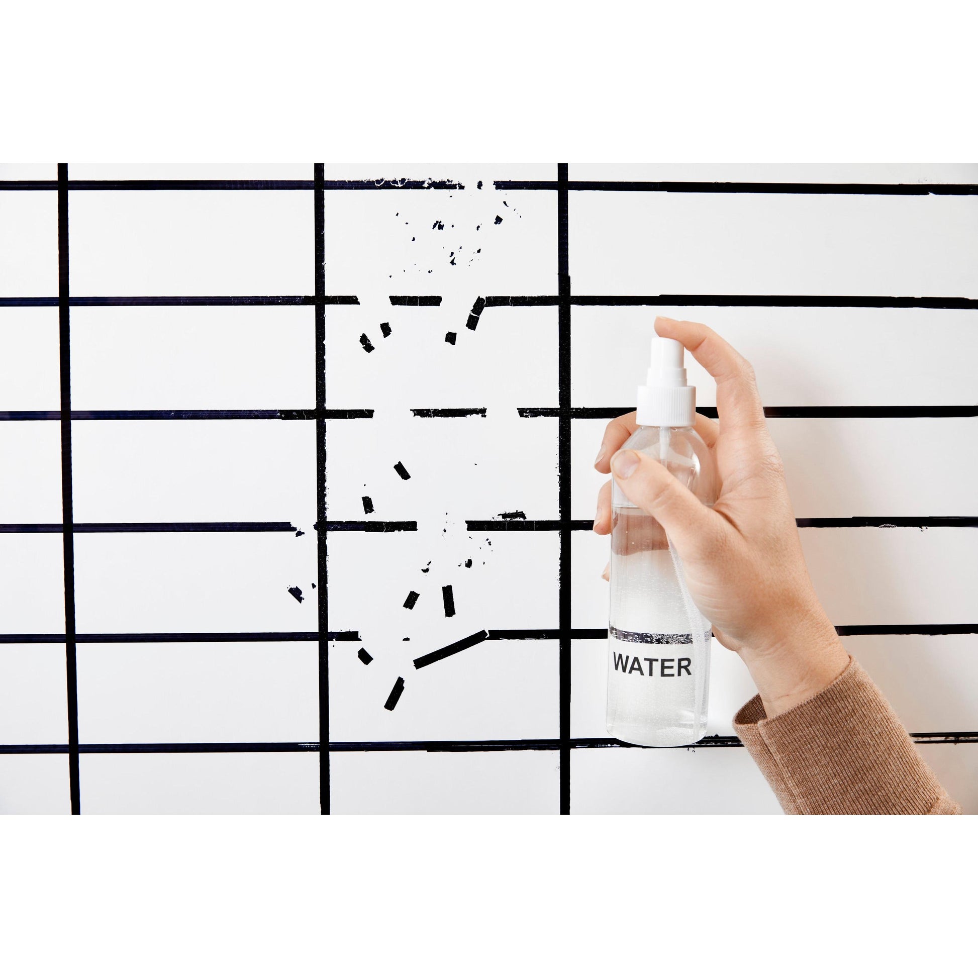 Eine Hand hält eine Sprühflasche mit der Aufschrift "WATER" vor einer Post-it® Easy Erase Whiteboard-Oberfläche, Weiß von 3M Deutschland GmbH, die ein schwarzes Gitter und einige fehlende oder gelöschte Linien aufweist.