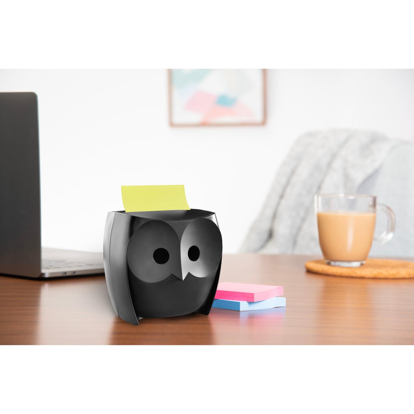 A 3M Deutschland GmbH Post-it® Z-Notes Dispenser, Owl, Black holds yellow notes, with pink and blue Super Sticky Z-Notes (45 sheets/block, 2 blocks/pack) lying next to it on a desk with a laptop, a coffee mug, and wall art in the background.