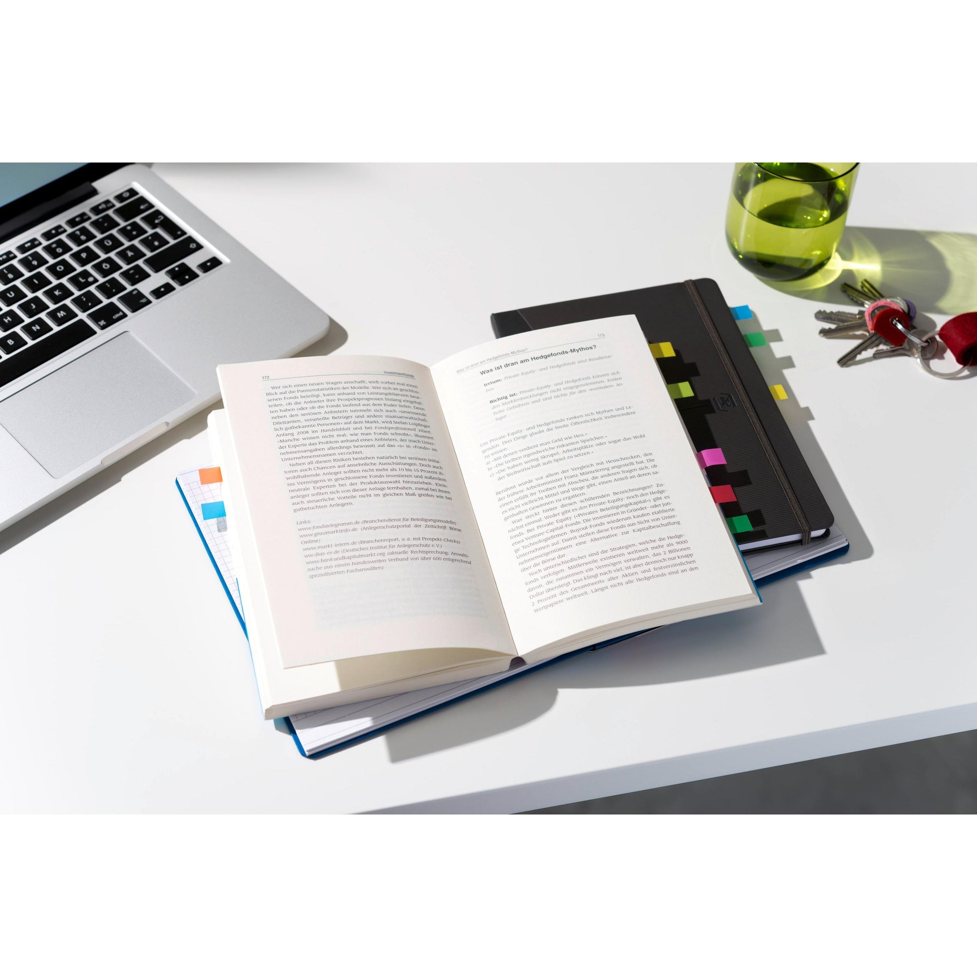 Ein offenes Buch liegt auf einem geschlossenen Notizbuch, das mit 3M Post-it® Index Haftstreifen Mini (11,9 x 43,2 mm) beschriftet ist. In der Nähe befinden sich ein Laptop, ein Glas grünes Getränk und Schlüssel mit einem roten Schlüsselbund auf dem weißen Schreibtisch.