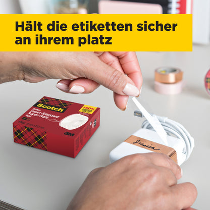 Eine Person wickelt ein weißes Etikett mit schwarzer Schrift um ein weißes Ladegerät, während auf dem Tisch eine Schachtel Scotch® Super-Hold Secure Tape, 1 Rolle, 19 mm x 25,4 m von 3M Deutschland GmbH steht. Im Hintergrund sind unscharfe Büromaterialien und eine gelbe deutsche Fahne zu sehen.
