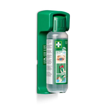 A 500 ml eye wash bottle from Orkla Wound Care AB is mounted on a wall holder (for eye wash 500 ml); a glow-in-the-dark instruction sign is included. The bottle displays illustrated instructions and an eye on the label.
