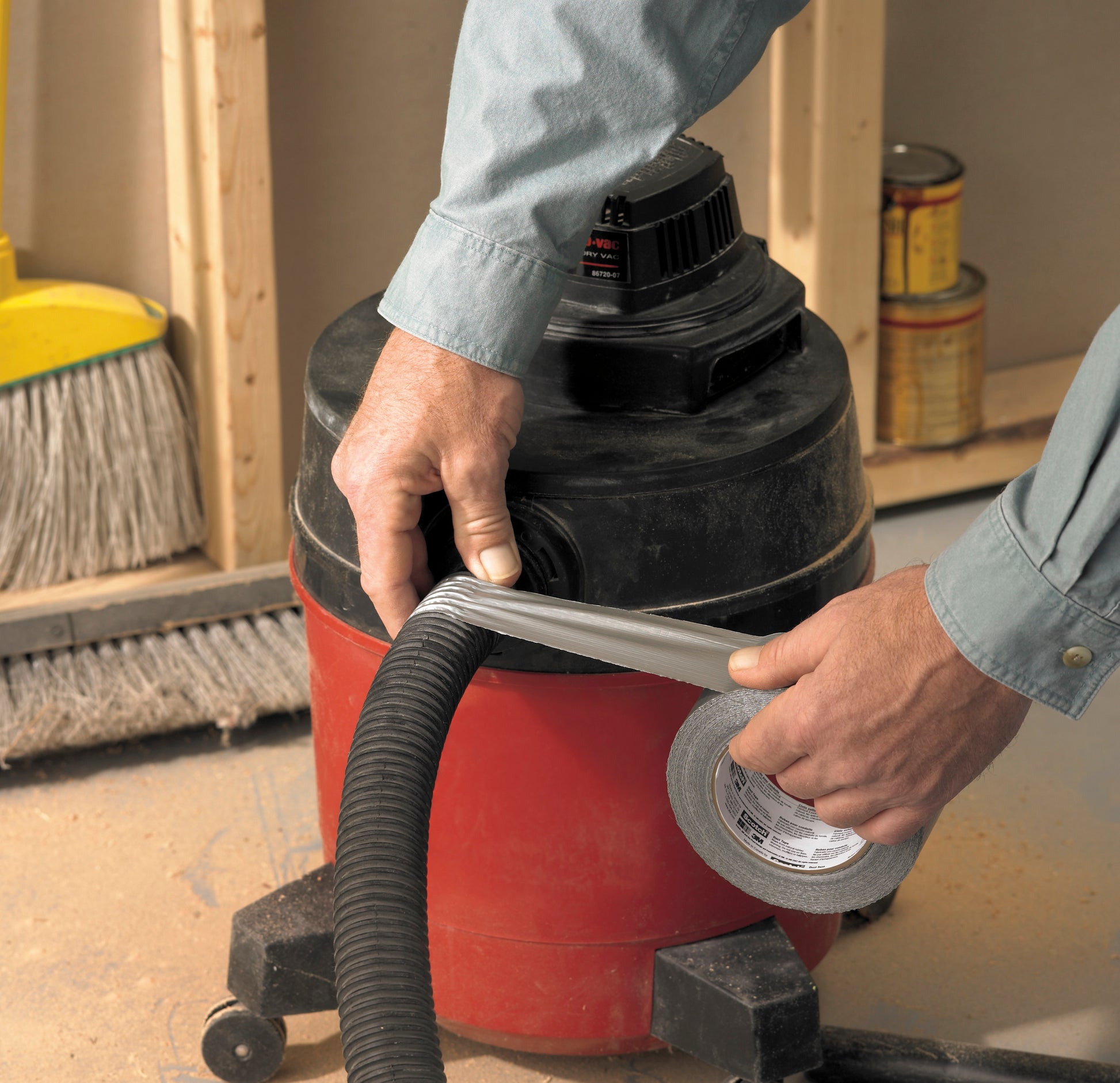 Jemand wickelt Scotch® Extremium™ Universal Klebeband (3 m x 19 mm) von 3M Deutschland GmbH um den Schlauch eines rot-schwarzen Werkstattsaugers. Ein Besen und Farbdosen liegen auf dem staubigen Boden einer Baustelle.