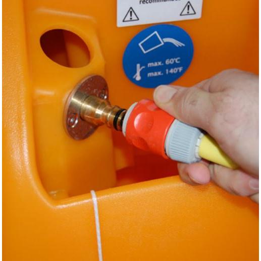 Eine Hand verbindet einen Schlauch mit einem roten und grauen AquaStop-Anschluss des TASKI swingo 2100 μicro von Diversey Deutschland GmbH & Co. OHG an eine Messingarmatur an einem orangefarbenen Behälter an, in dessen Nähe sich Warn- und Hinweisschilder befinden.