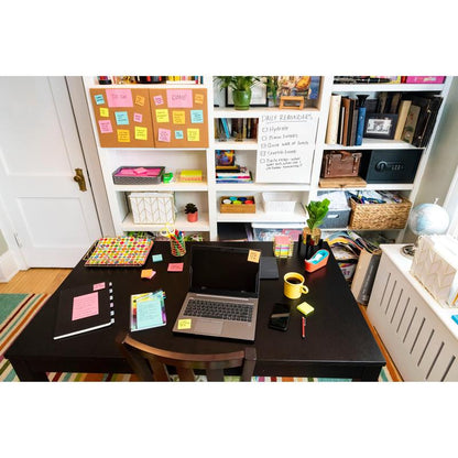 A desk with a laptop, notebook, phone, and 3M Deutschland GmbH Post-it® Super Sticky Notes (76 mm x 76 mm, 90 sheets/block, 100% PEFC) stands in front of a bookshelf with books and a whiteboard for daily reminders; colorful materials ensure organization.
